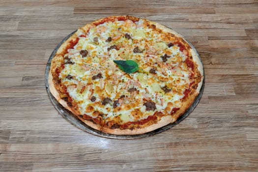 A pizza with a green leaf on top sits on a wooden table. The pizza is topped with cheese and meat, and it looks delicious