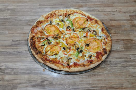 A pizza with tomatoes, mushrooms, and peppers sits on a wooden table. The pizza is cut into slices and ready to be eaten
