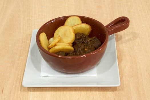 A bowl of food with potatoes and meat in it. The bowl is on a white plate. The plate is on a wooden table