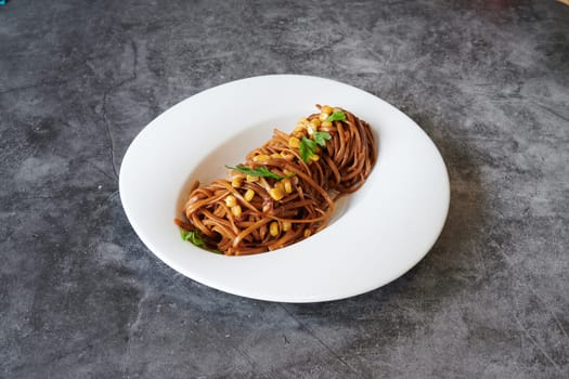 A plate of spaghetti with meat and vegetables. The plate is white and the spaghetti is brown. The plate is on a grey surface