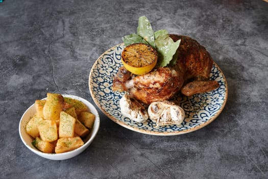 A plate of chicken with potatoes and mushrooms on top. The plate is blue and white. The chicken is cooked and has a lemon on top. The potatoes are in a bowl