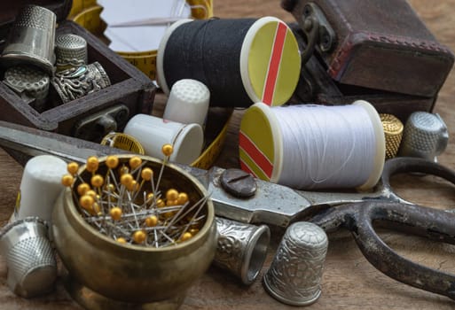Sewing needles stuck in a white paper, Thimbles in small wooden treasure chest, Spools of black and white thread, Steel scissors and Measuring tape including pins in brass cup on wooden table. Sewing items set or Sewing objects (Sewing notions), use it as your Wallpaper, Poster and Space for text, Selective focus.