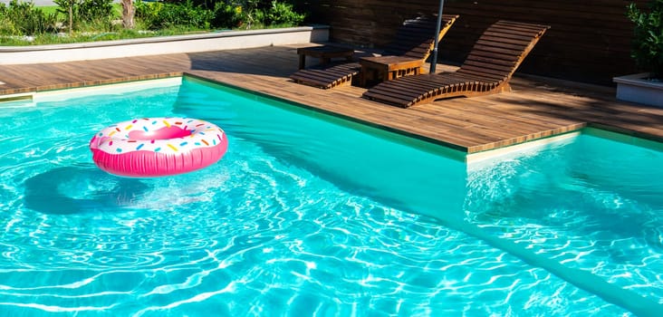 Turquoise swimming pool with pink inflatable ring, wooden deck, and modern lounge chairs in sunny backyard