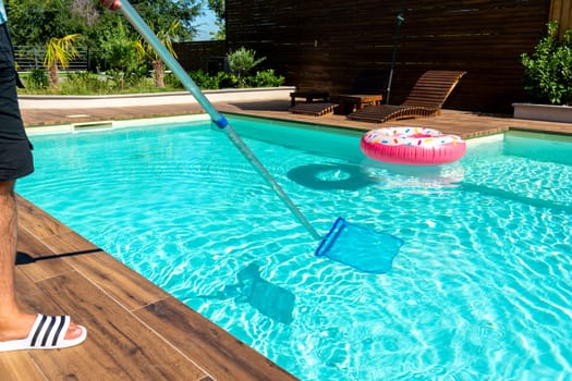 Man cleaning turquoise swimming pool water with skimmer net, wooden deck, and colorful inflatable floats