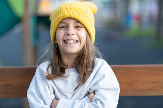 Portrait of cheerful pretty girl in yellow cap with freckles on face looking at camera while sitting on bench