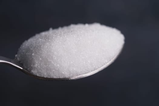 A spoonful of granulated sugar is displayed against a dark backdrop.