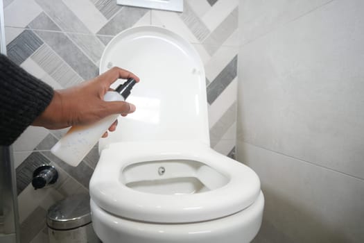 A person cleans a toilet using a spray bottle in a tidy bathroom.