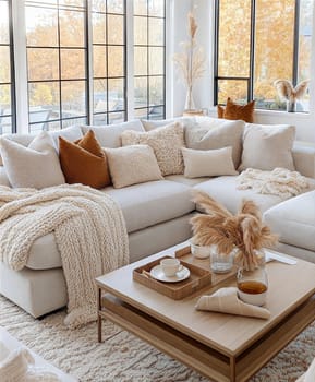 A stylish and cozy minimalist living room featuring a soft white sofa, wooden coffee table, fluffy pillows, knitted blanket, books, ceramic cups, and dried flowers in a serene neutral color palette