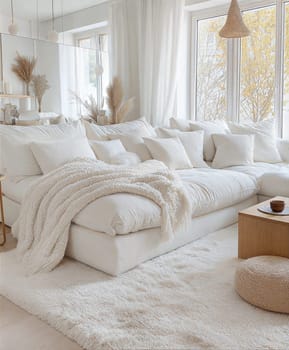 A stylish and cozy minimalist living room featuring a soft white sofa, wooden coffee table, fluffy pillows, knitted blanket, books, ceramic cups, and dried flowers in a serene neutral color palette