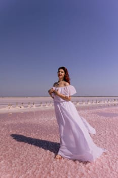 A woman in a white dress stands on a pink salt flat. The sky is blue and there is no wind. The woman is looking to the left.