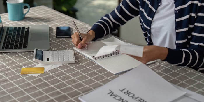 A person is focused on taking notes for financial planning and bills, using a calculator and holding documents, with a notebook open on the table.