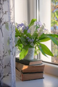 beautiful bouquet of lily of the valley and blue phlox, standing in decorative vase on stack of old books near window with sheer curtain,cozy and peaceful atmosphere in morning sunlight