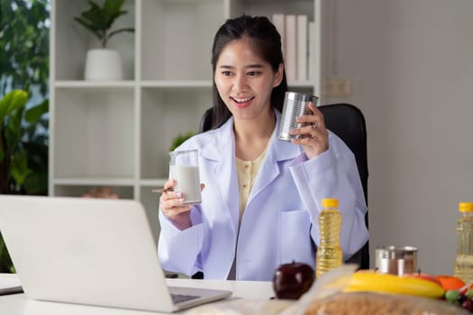 A nutritionist is showcasing healthy food options, holding a glass of milk and a can, while interacting with clients via a laptop.