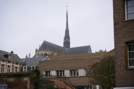 The towering spire of Amiens Cathedral rises above surrounding brick buildings, showcasing its Gothic architecture on a cloudy day.
