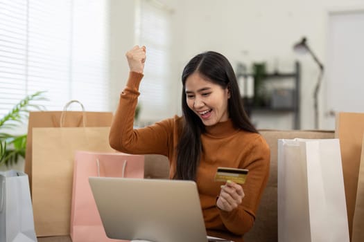 An excited woman raises her fist in celebration while shopping online with her credit card and laptop, surrounded by shopping bags.