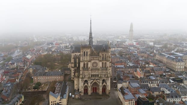 Aerial view of the Gothic Amiens Cathedral in northern France, surrounded by a foggy urban cityscape. This UNESCO site showcases stunning medieval architecture in a historic and atmospheric setting
