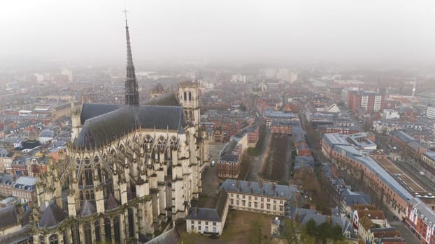 Aerial view of the iconic Gothic cathedral in Amiens, France, surrounded by urban cityscape under a misty, overcast sky.