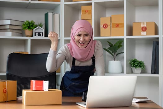 A delighted Muslim woman raises her fist in excitement while working on her online business in a cozy office setting.