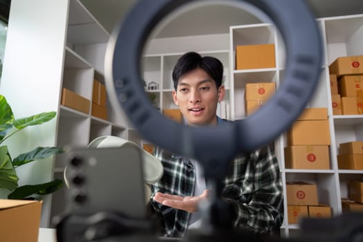 A young entrepreneur is presenting a product online, utilizing a smartphone and ring light in a modern workspace filled with product boxes.
