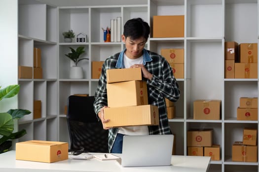 A young entrepreneur is organizing product boxes while overseeing online sales, working in a modern workspace with a laptop.