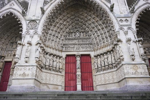 Detailed view of the Amiens Cathedral entrance in France, featuring intricate stone carvings, statues, pointed arches, and vibrant red doors.