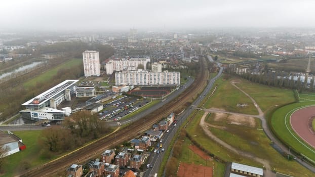 Aerial view of a neighborhood in Amiens, France, featuring residential buildings, roads, railway tracks, and green fields. A blend of urban planning and natural landscapes.