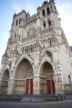 Majestic facade of a Gothic cathedral showcasing intricate sculptures, pointed arches, and vibrant red doors against a blue sky. Amiens. France 17.02.2025