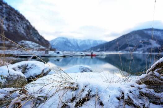 Close-up of snow-covered grass with a tranquil marina, calm fjord, and snowy mountains in the background in Gaupne, Norway.