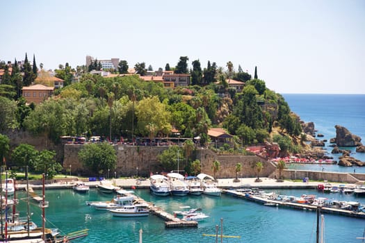 A picturesque view of Antalya marina, featuring boats docked along turquoise waters, historic stone walls, lush greenery, Mediterranean houses, and