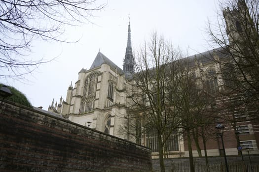 Majestic Gothic cathedral with intricate architecture and a tall spire, framed by bare trees and a stone wall in Amiens, France