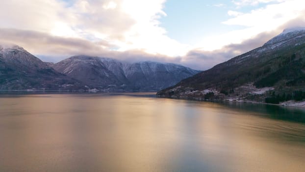 Aerial winter view of Gaupne, Norway, featuring a fjord reflecting golden sunset light, surrounded by snow-covered mountains and a peaceful rural landscape.