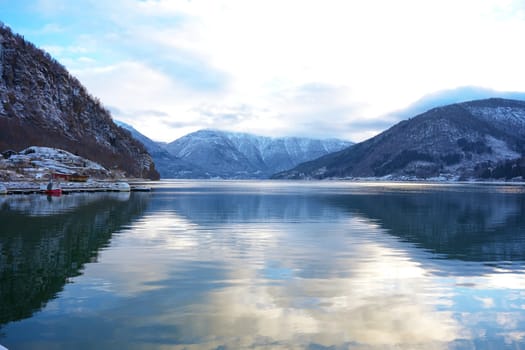 A serene fjord surrounded by snow-covered mountains reflects the peaceful winter scenery in Gaupne, Norway, under soft light.