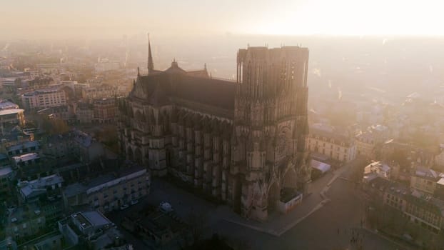 Aerial view of Reims Cathedral during sunrise, bathed in golden light. The Gothic landmark overlooks the historic cityscape of Reims, France, in a serene atmosphere.
