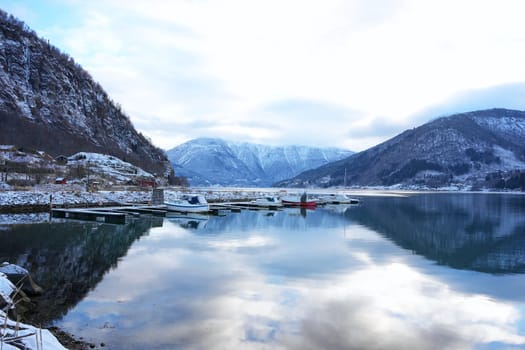 A serene marina in Gaupne, Norway, featuring boats docked on calm water, surrounded by snowy mountains and beautiful reflections.