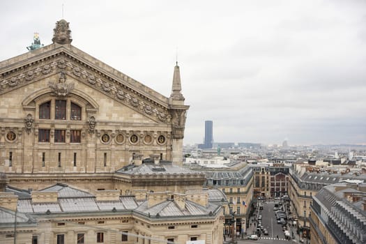 A close-up of the intricate facade of the Opera Garnier in Paris, with classic rooftops, bustling streets, and a modern skyscraper in the background under cloudy skies.