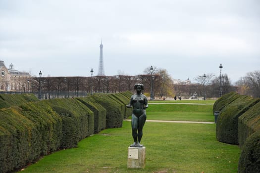 A serene view of the Tuileries Garden featuring a bronze statue, neatly trimmed hedges, and the Eiffel Tower under overcast skies. Paris, France 20.01.202