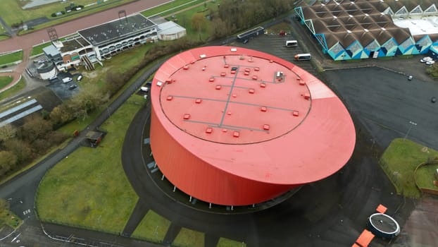 Aerial view of a striking zenith arena building featuring a modern architectural design. Surrounded by green spaces, parking, and adjacent facilities, it stands out as a unique structure in the area.