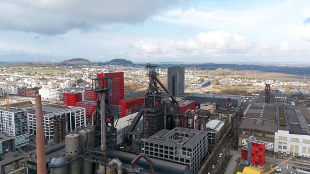 A high-angle aerial view of Esch-sur-Alzette city, showcasing a blend of modern architecture, historic steel industry structures, and surrounding residential and natural landscapes.