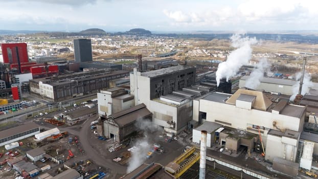 High-angle aerial view of Esch-sur-Alzette, Luxembourg, showcasing a large steel factory with smokestacks emitting steam, surrounded by industrial buildings, warehouses, and modern urban developments.
