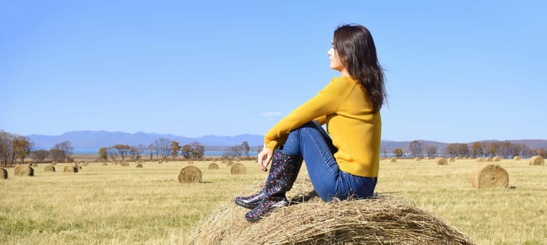 Young brunette woman in yellow sweater and rubber boots sitting on top of haystack in field. she looking at mountains, Japanese sea and the autumn forest. Primorsky kray, Russia. Beauty in nature.