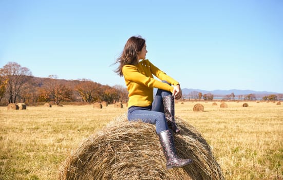 Young brunette woman in yellow sweater and rubber boots sitting on top of haystack in field. she looking at mountains, Japanese sea and the autumn forest. Primorsky kray, Russia. Beauty in nature.
