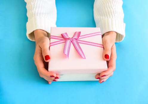 Pink gift box with bow in female hands in white sweater, with red nail polish, on a blue background.