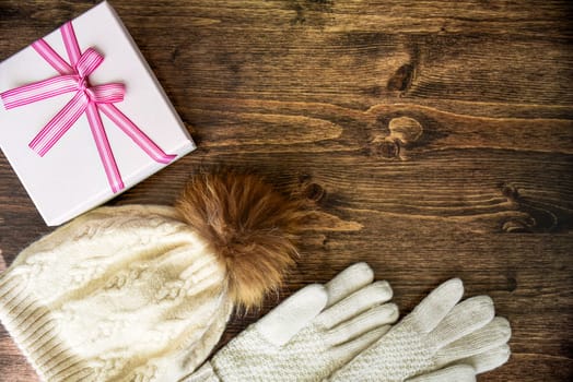 Wooden background with warm white hat with pom pom, gloves and gift box with pink bow. Christmas and winter mood, copy space.
