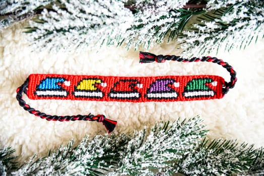 Woven friendship bracelet with bright alpha pattern Multicolored christmas hats, handmade of thread, next to snowy fir branches.