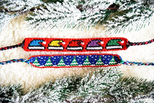 Woven friendship bracelets with alpha patterns Snowy fir trees, Santa hats. Handmade of thread on light background with  snowy branches. DIY New Year gift idea