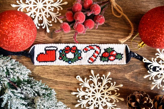 Woven friendship bracelet with pattern Christmas berry, lollipop, wreath and santas boot. Handmade of thread, on wooden background with snowflakes, spruce cone,