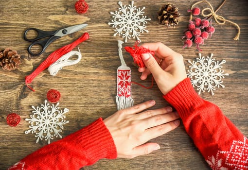Process of weaving friendship bracelet from floss threads.female hands in red sweater braiding bracelet. scissors, embroidery thread,fir cones and berries on wooden background.