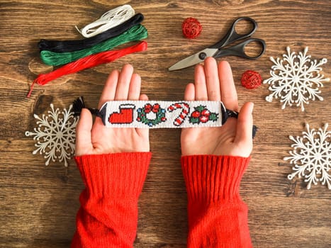 Idea for Christmas gift. Friendship bracelet with New Year alpha pattern handmade of floss threads in female hands in red sweater. Scissors, skeins of embroidery thread on wooden background.