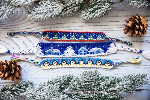 Woven friendship bracelets with alpha patterns Snowy night with houses,penguins, fir trees. Handmade of thread, on wooden background with spruce cones, snowy branches.