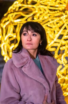 Russian 48 years old woman in blue sweater and brown coat standing against yellow christmas lights before Christmas